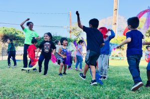A group of young children and an adult play energetically on a grassy field in Santa Barbara County, waving colorful scarves and smiling under a clear blue sky. The scene is lively and joyful, with everyone enjoying the outdoor activity.