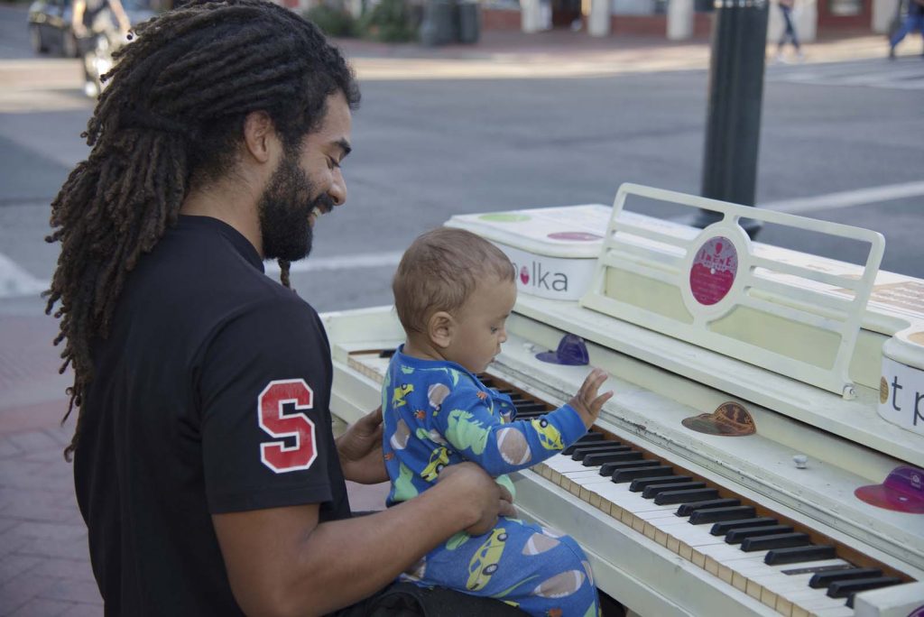 A man with dreadlocks smiles while holding a baby in pajamas on his lap as they play a white outdoor piano together on a Santa Barbara County city sidewalk.