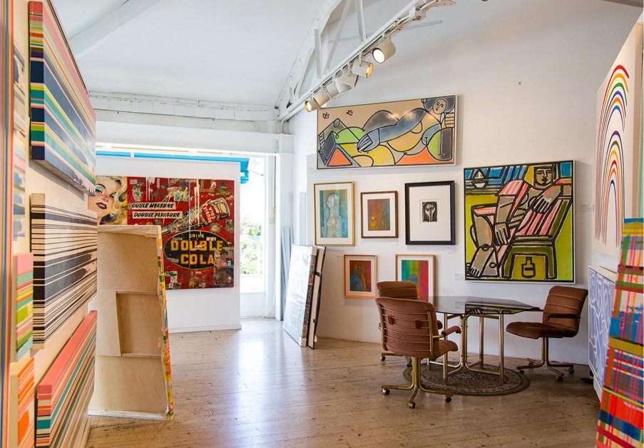 A brightly lit art gallery in Santa Barbara County with colorful abstract and pop art paintings on white walls, a round table with two chairs, and wooden floors. Framed artwork is arranged closely together throughout the space.