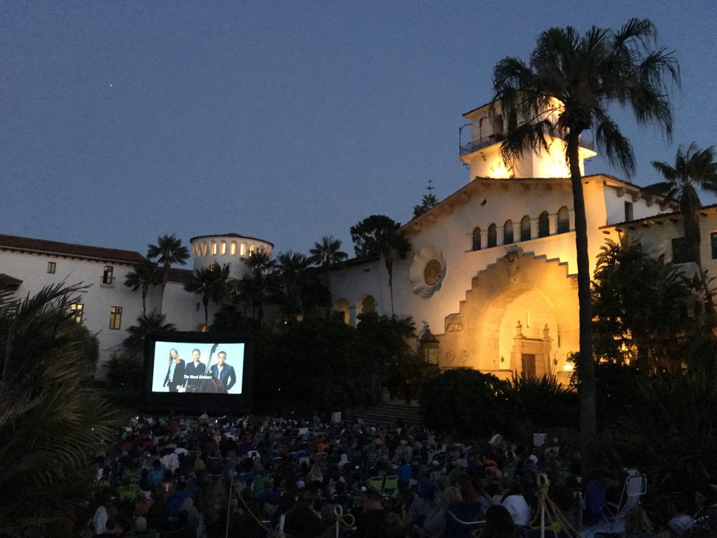 A large crowd sits outdoors at dusk in Santa Barbara County, watching a movie projected on a screen in front of an illuminated Spanish-style building surrounded by palm trees.