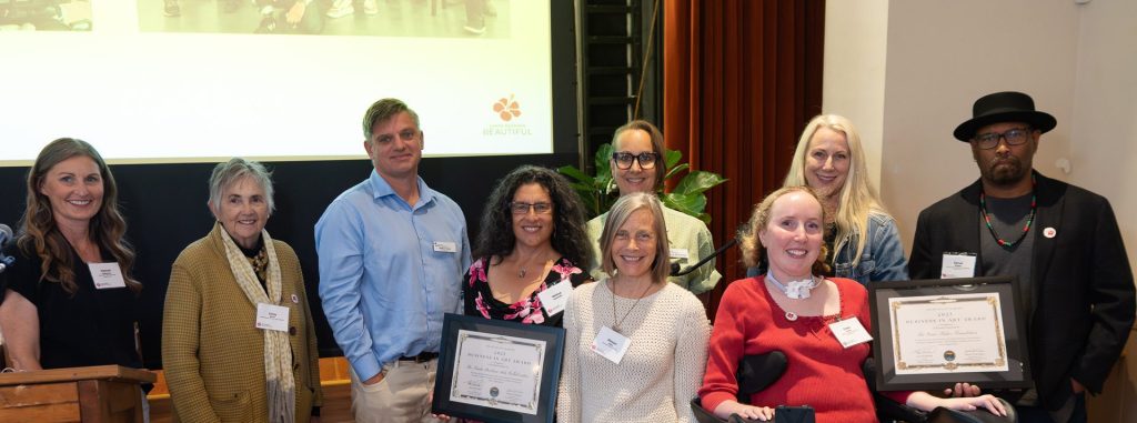 A group of nine people, some holding certificates, pose and smile together indoors at an event. One woman in a wheelchair holds a certificate. There is a wooden podium, green plants, and a projected image in the background.