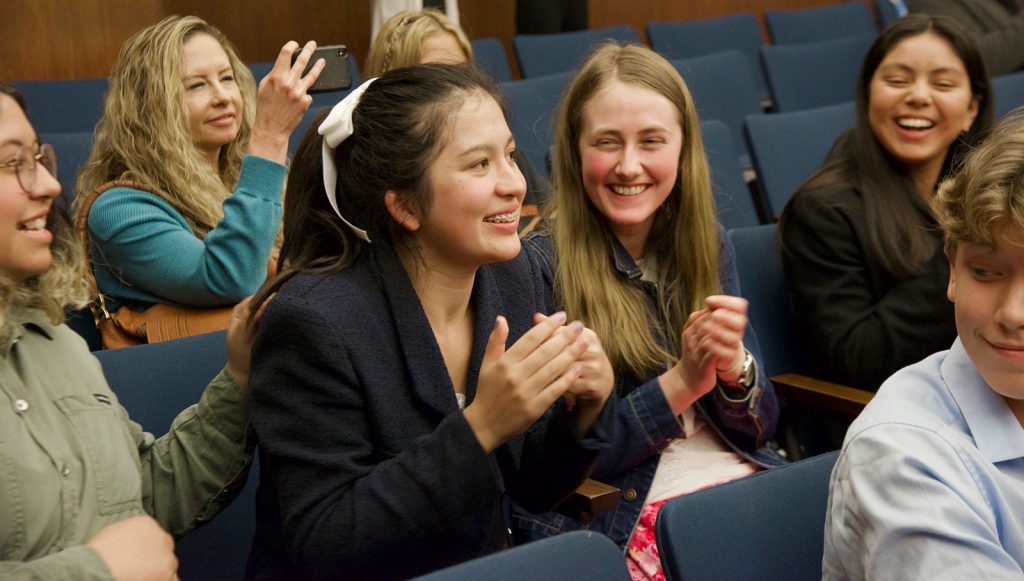 A group of young people in Santa Barbara County sit in blue auditorium chairs, smiling and clapping. One person in front is clapping enthusiastically, while others around her laugh and cheer. A woman in the back snaps a photo with her phone.