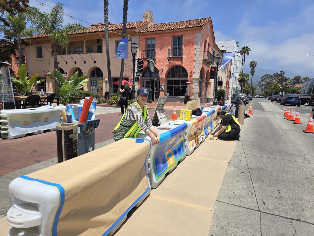 Three people in safety vests decorate street barriers with colorful designs on a Santa Barbara County sidewalk, working near orange cones and a pink stucco building lined with palm trees.