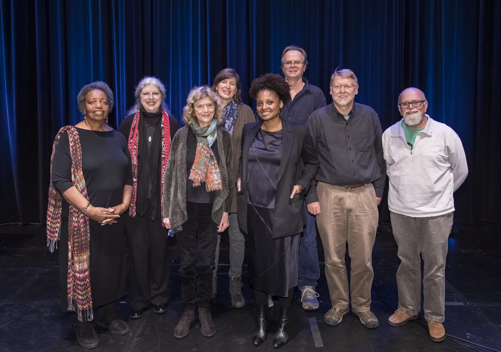 A group of nine adults, dressed in casual and semi-formal clothing, stand together on a stage with a black floor and dark blue curtains in the background. They are smiling and posing for the photo at a Santa Barbara County event.