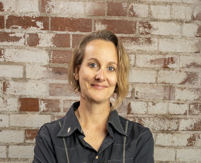 A woman with short blonde hair, wearing a dark button-up shirt, stands in front of a light-colored brick wall in Santa Barbara County and smiles with her arms crossed.