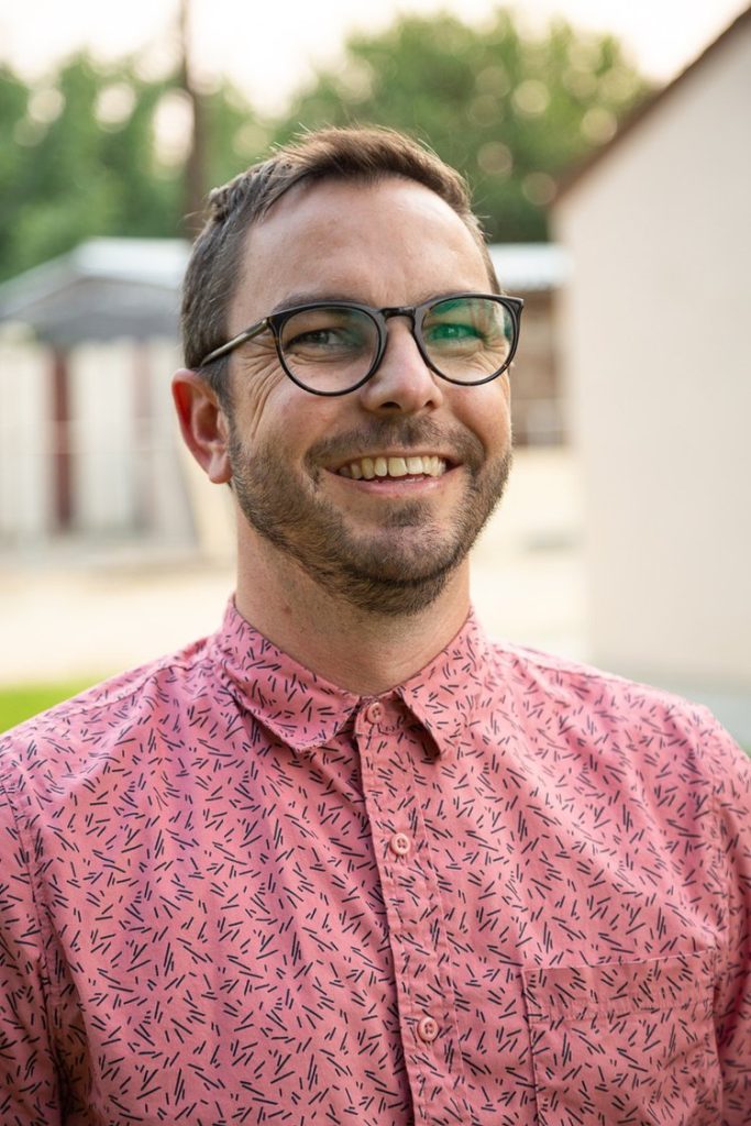 A smiling man with short brown hair, a trimmed beard, and glasses wears a pink patterned button-up shirt. He stands outdoors in Santa Barbara County, with blurry greenery and buildings in the background.