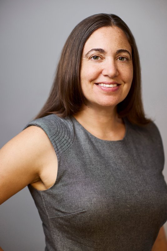 A woman with long brown hair and a gray sleeveless top smiles at the camera against a plain gray background, embodying the relaxed charm of Santa Barbara County.