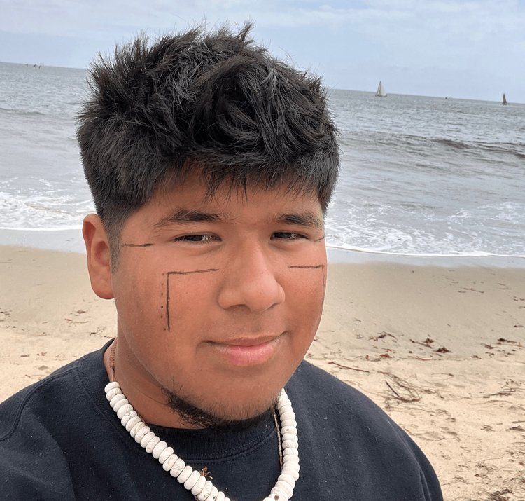 A young person with short, dark hair and geometric face paint smiles at the camera on a sandy Santa Barbara County beach, wearing a black shirt and a white shell necklace. The ocean and sailboats are visible in the background under a cloudy sky.