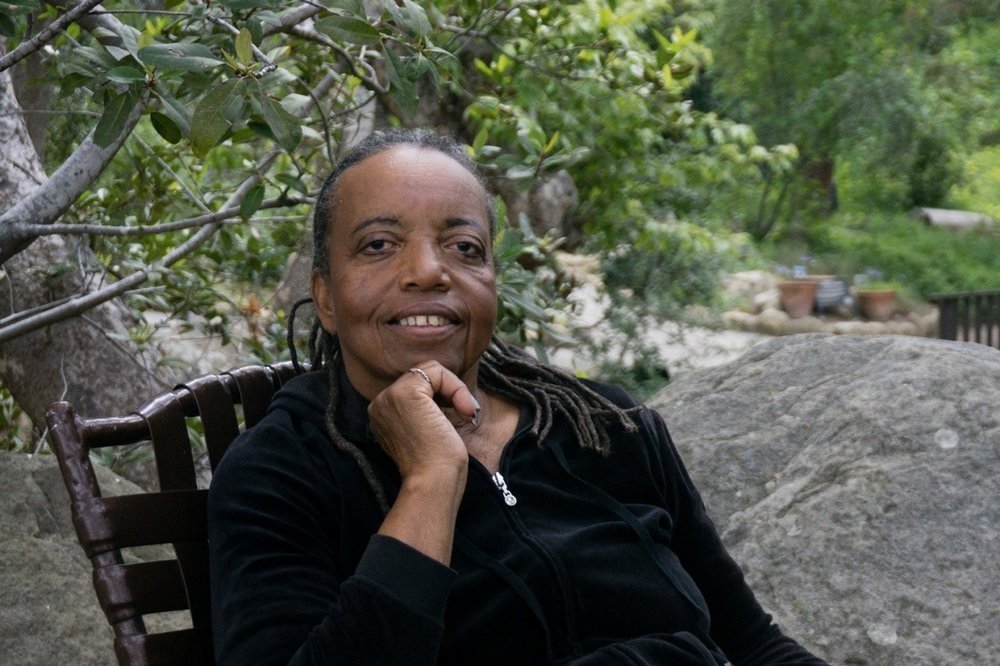 A woman with short, dark hair and a black zip-up jacket sits outdoors on a wooden chair in Santa Barbara County, smiling and resting her chin on her hand, with trees and large rocks in the background.