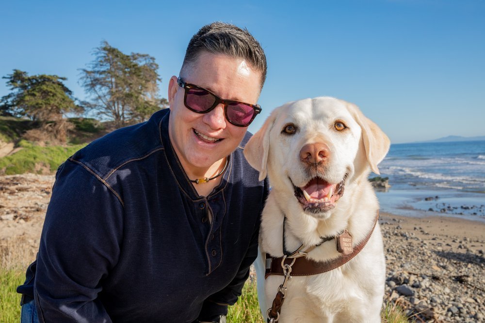 A person with short hair and sunglasses smiles next to a yellow Labrador wearing a harness on a sunny Santa Barbara County beach, with trees and the ocean in the background.
