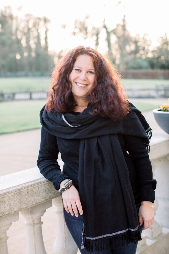 A woman with long, wavy, reddish-brown hair smiles while standing outdoors by a stone railing in Santa Barbara County. She is wearing a black sweater, black scarf, and bracelets, with trees and a lawn in the background.