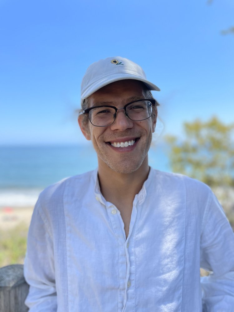 A person wearing glasses, a white cap, and a white shirt smiles while standing outdoors in Santa Barbara County, with the ocean and blue sky in the background. Trees and a sandy beach are visible behind them.