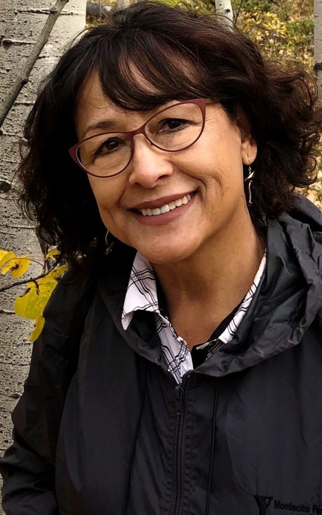 A woman with short dark curly hair, glasses, and a black jacket smiles outdoors in Santa Barbara County, framed by tree trunks and autumn leaves.