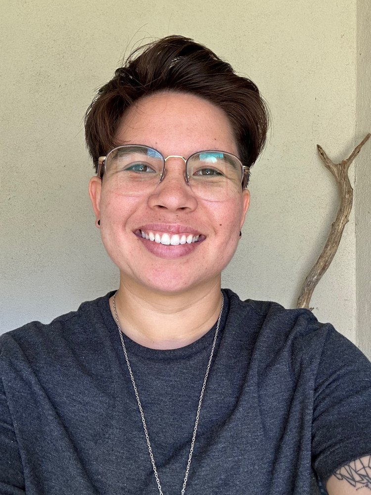 A person with short brown hair, glasses, and a dark t-shirt smiles at the camera. They are indoors in Santa Barbara County with a plain light-colored background and a branch visible on the right side.
