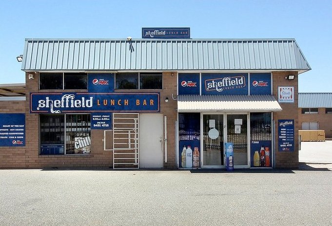 A brick building with blue signs reading Sheffield Lunch Bar above the entrance and windows. The windows display beverage ads, and the glass doors have an Open sign. The area in front is empty and sunny.