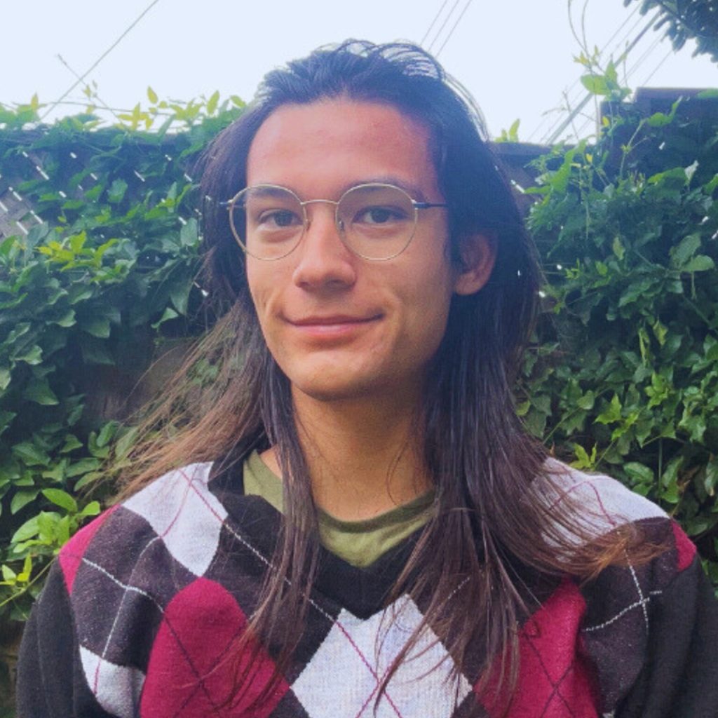 A person with long brown hair and glasses smiles slightly while standing outside in front of green leafy vines. They are wearing a patterned sweater with red, black, and white diamonds over a green shirt.