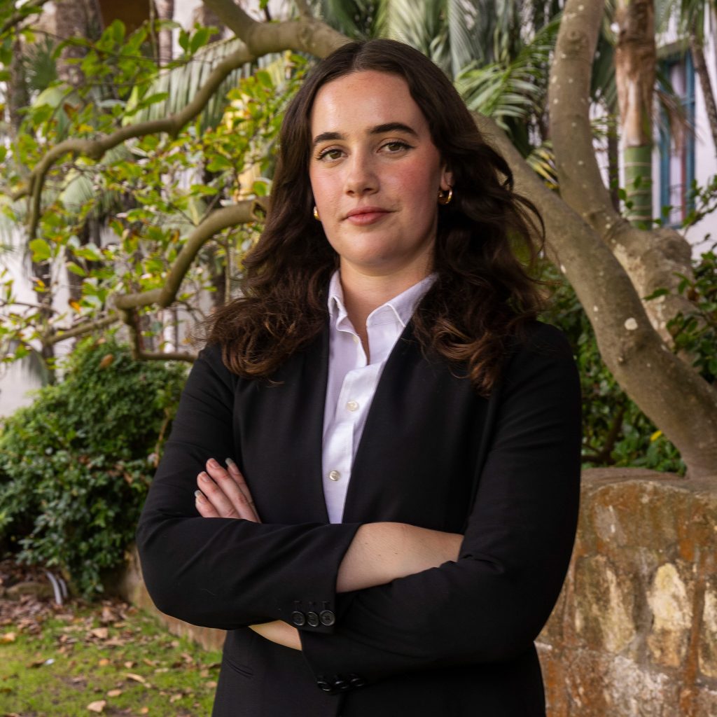 A woman with long dark hair, wearing a black blazer and white shirt, stands outdoors with arms crossed in front of greenery and a stone wall. She has a confident expression.