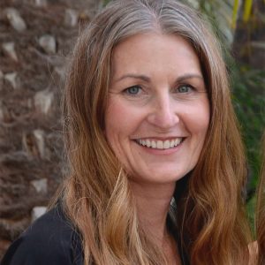 A woman with long, wavy light brown hair smiles at the camera. She is outdoors, with a blurred background of stone and greenery. She is wearing a dark top.