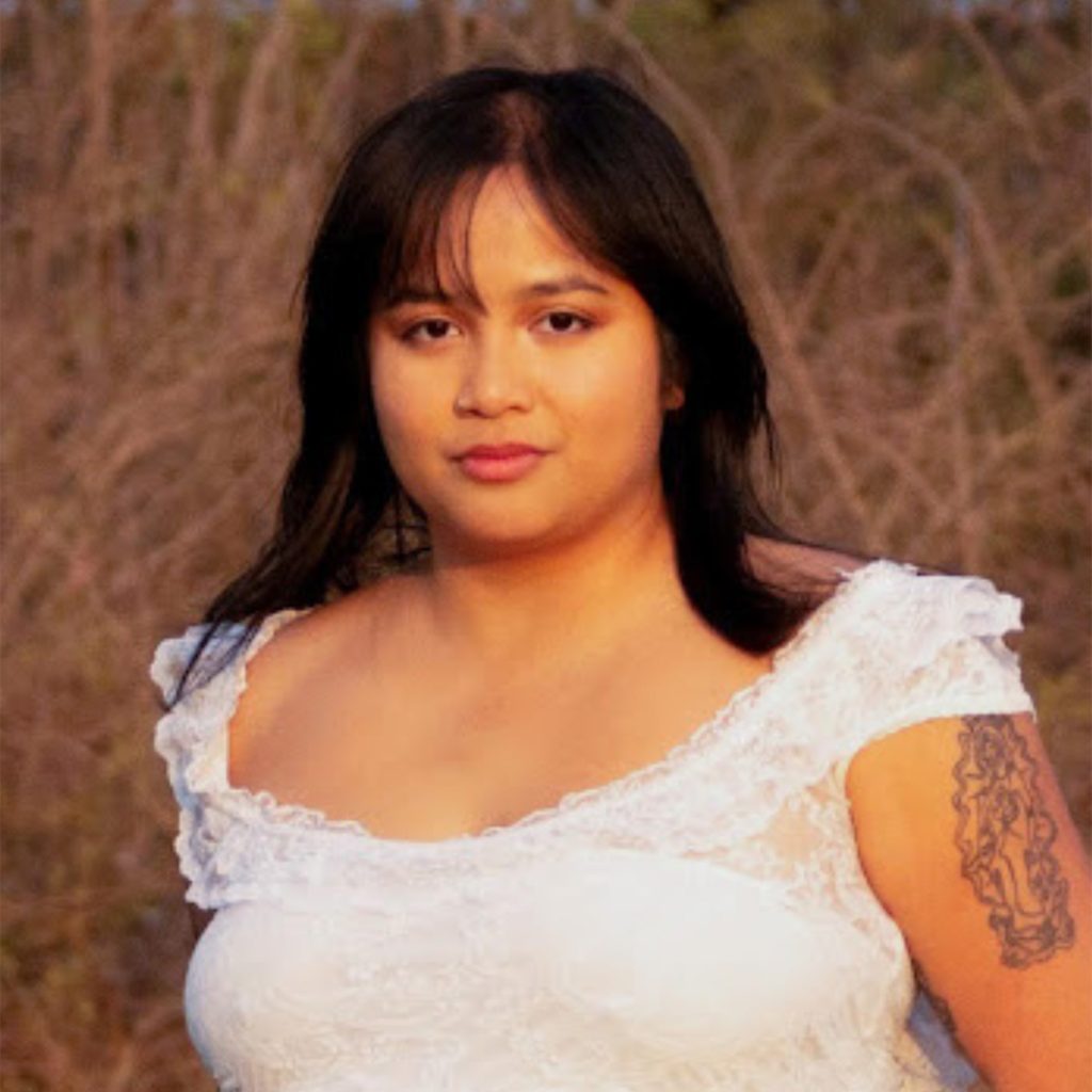A person with medium-length dark hair, wearing a white lace dress, stands outdoors in front of tall, dry grass. They have a tattoo on their upper arm and a calm expression.