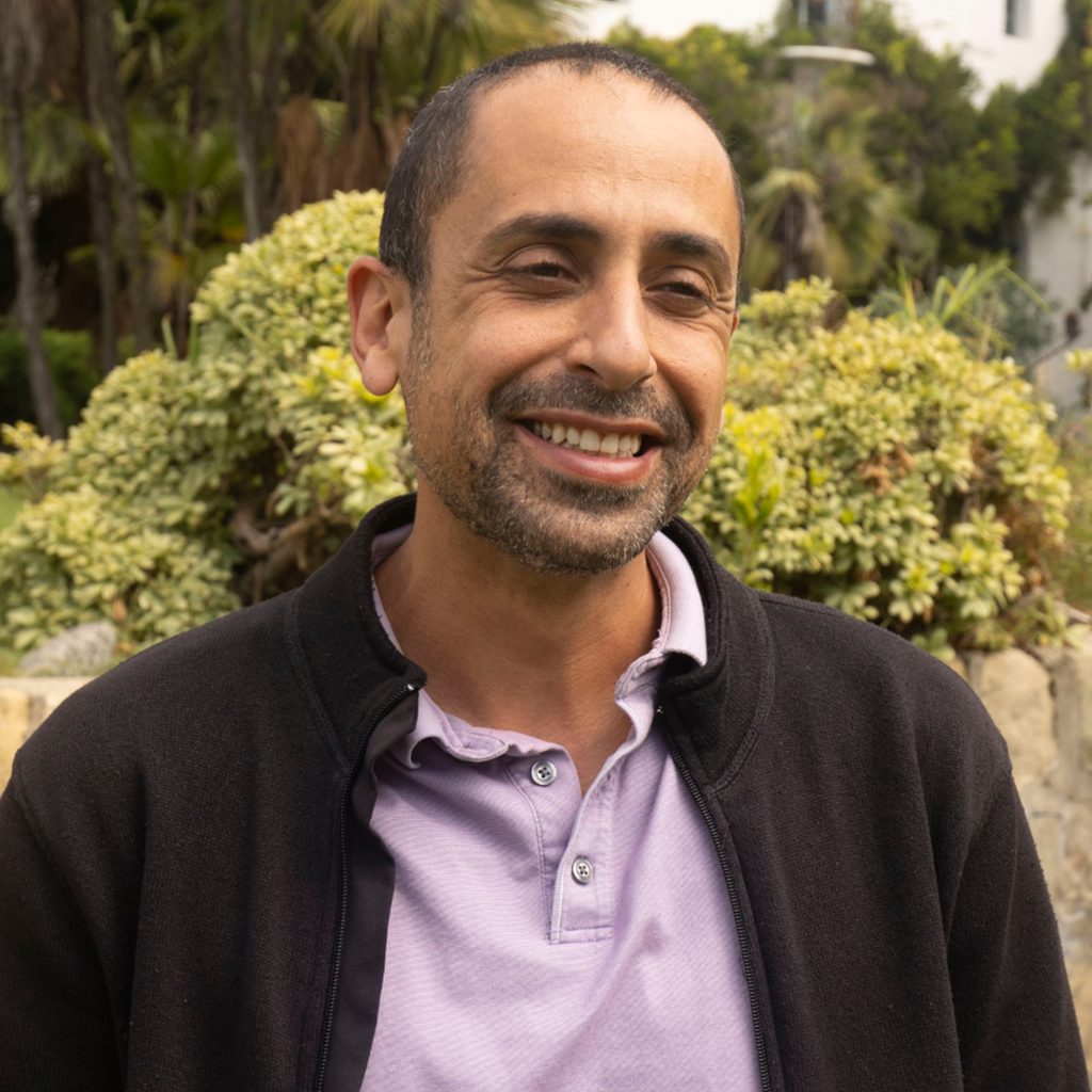 A man with short dark hair and a trimmed beard, wearing a light purple polo shirt and black jacket, smiling outdoors in front of green bushes.