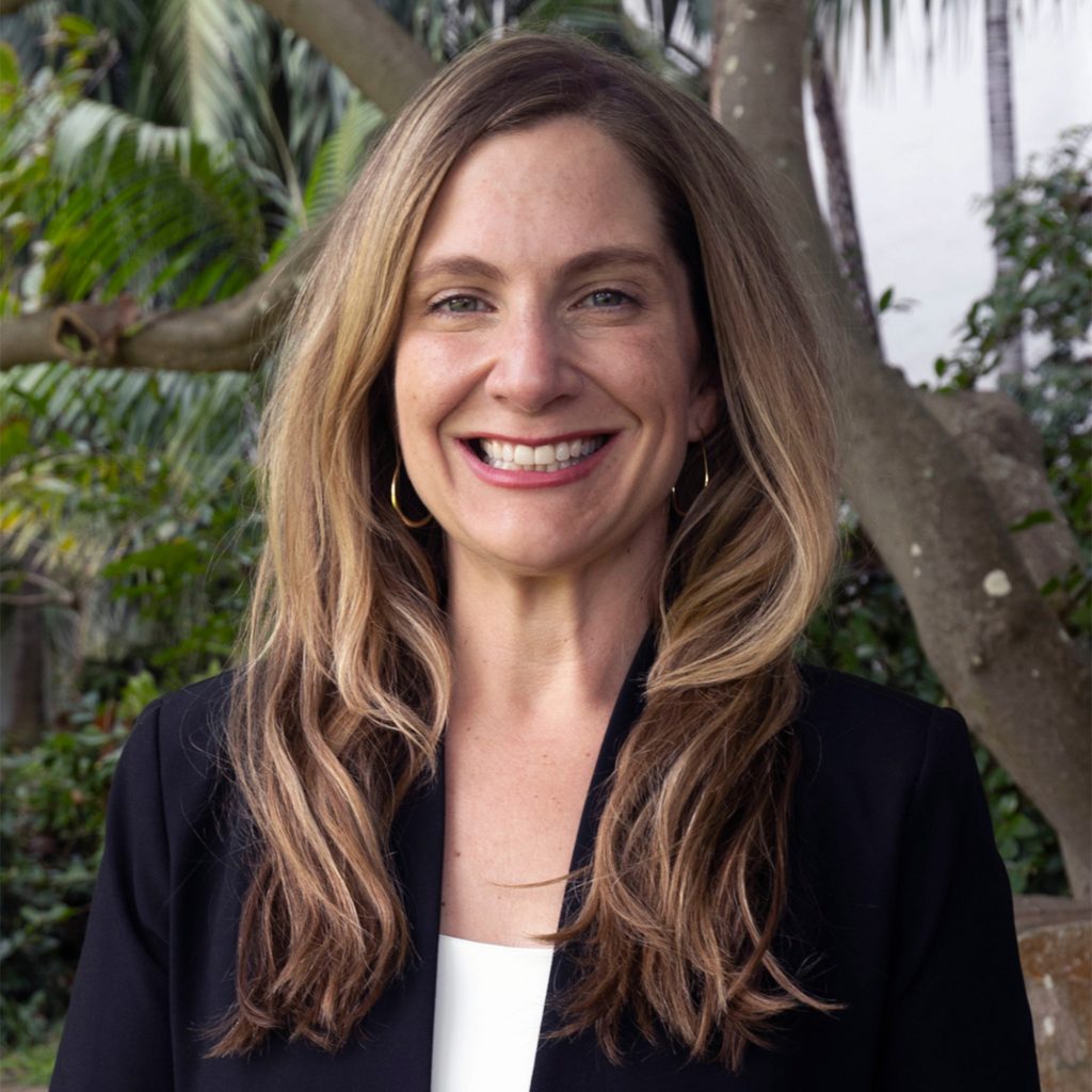 A woman with long, wavy light brown hair is smiling and standing outdoors. She is wearing a black blazer over a white top, with gold hoop earrings. Green foliage and tree branches are visible in the background.