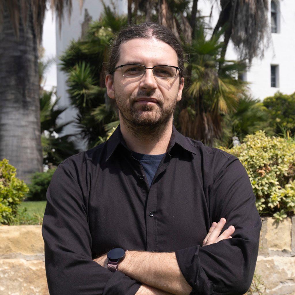 A man with long hair tied back, glasses, and a beard stands outdoors with arms crossed, wearing a black shirt and watch. Behind him are palm trees, hedges, and a white building.