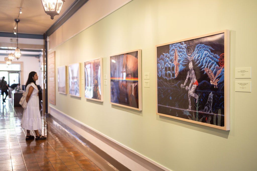 A woman in a white dress stands in an art gallery, looking at a series of colorful framed paintings displayed on a long, light-green wall under hanging lights.