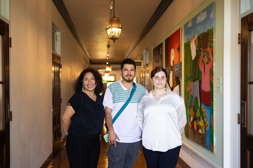 Three people stand smiling in a well-lit hallway lined with colorful paintings on the wall, wood doors on either side, and hanging lantern-style lights above.