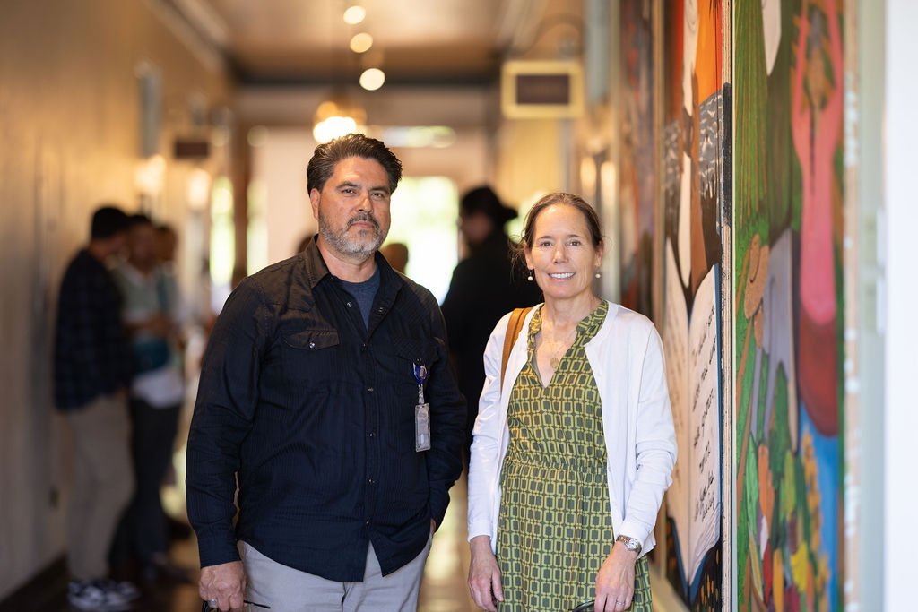 A man and a woman stand side by side in a well-lit hallway with colorful artwork on the walls. Several people are blurred in the background, suggesting a social or professional event.