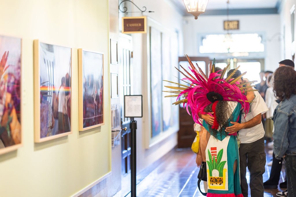 A person wearing a colorful traditional costume with a large feathered headdress stands in a hallway lined with artwork, as people observe the scene.