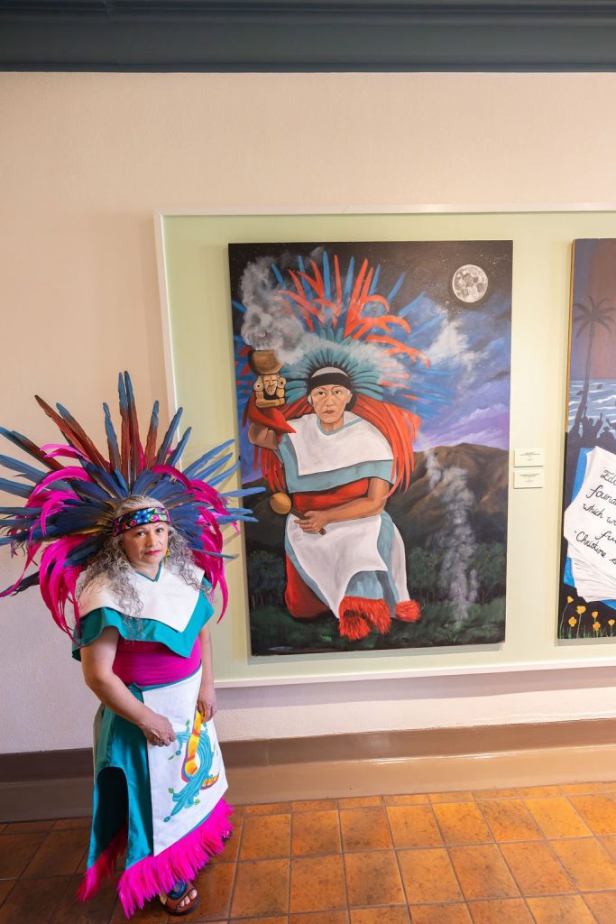 A person wearing a colorful feathered headdress and traditional clothing stands next to a painting of an Indigenous figure in similar attire, holding a cup with a night sky in the background.