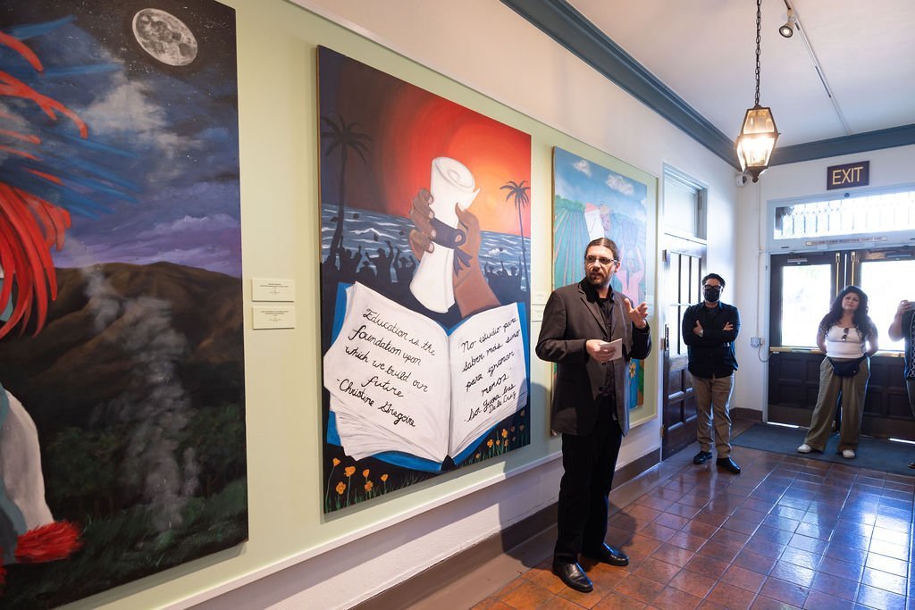 A man in a suit gestures while speaking to a small group in a hallway with large, colorful paintings on the wall. Three people stand near the door, listening. The space is well-lit with natural light.