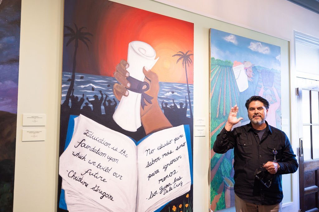 A man stands indoors, waving and holding glasses in front of a colorful mural showing a hand holding a diploma by the ocean, an open book with handwritten quotes, and people raising their arms in celebration.