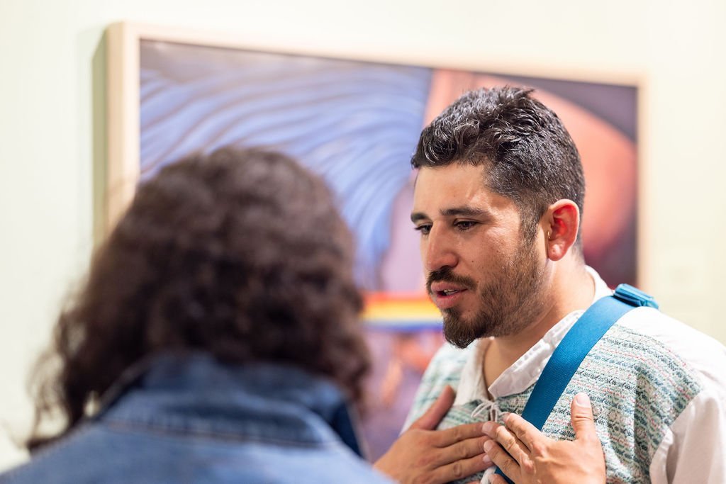 A man with dark hair and a trimmed beard gestures with his hands on his chest while speaking to another person with long hair. They are standing in front of a blurred painting in a gallery or museum.