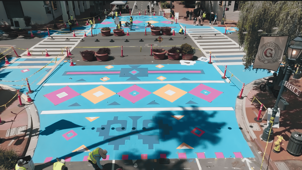 A city street painted with vibrant geometric patterns in blue, pink, orange, and yellow. Workers in safety vests are painting, with traffic cones and planters blocking off the area. A large tree shadow falls over the mural.