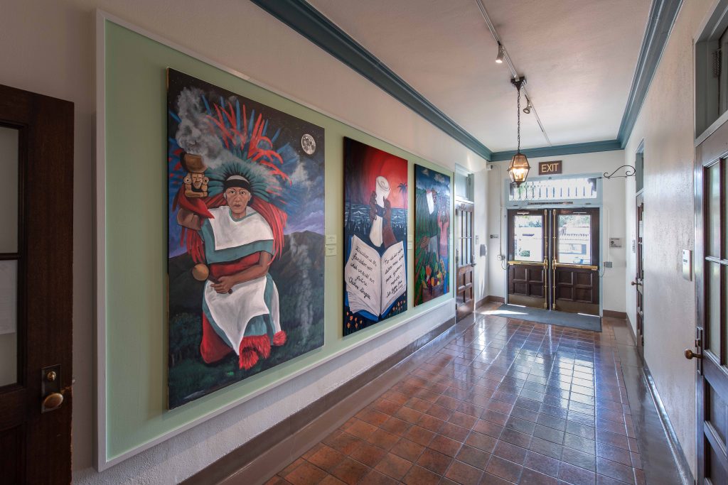 A hallway with brown tile floor and light walls features three large colorful paintings, including a woman in white, a book with stars, and a red sunset. The hallway has an exit door at the end and hanging light fixtures.