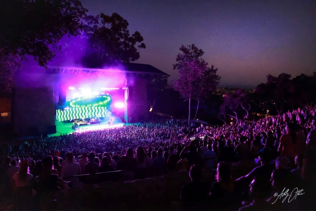 An outdoor concert at sunset, with a large crowd seated facing a brightly lit stage. Colorful lights illuminate the performers and the surrounding trees, creating a vibrant, energetic atmosphere.